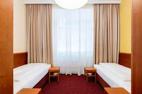 Zwei Einzelbetten stehen in einem modernen, hellen Zimmer mit einem großen Fenster. Vor dem Fenster hängen Vorhänge, und es gibt kleine Holztische neben den Betten. Der Teppichboden ist rot.