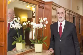 Ein älterer Mann mit Brille und grauem Bart trägt einen schwarzen Anzug und eine rote Krawatte. Er steht neben einem Tisch mit einer gelben Blume. Im Hintergrund spiegelt sich ein heller Raum mit Holzvertäfelung und weiteren Pflanzen.