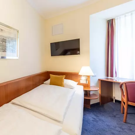 Ein gemütliches Hotelzimmer mit zwei Einzelbetten und einem gelben Kissen. An der Wand hängt ein gerahmtes Bild. Ein Fernseher ist über den Betten montiert. Neben einem Fenster ist ein kleiner Tisch mit einem Stuhl, während eine Lampe auf einem Nachttisch leuchtet.
