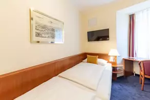 Ein gemütliches Hotelzimmer mit zwei Einzelbetten und einem gelben Kissen. An der Wand hängt ein gerahmtes Bild. Ein Fernseher ist über den Betten montiert. Neben einem Fenster ist ein kleiner Tisch mit einem Stuhl, während eine Lampe auf einem Nachttisch leuchtet.