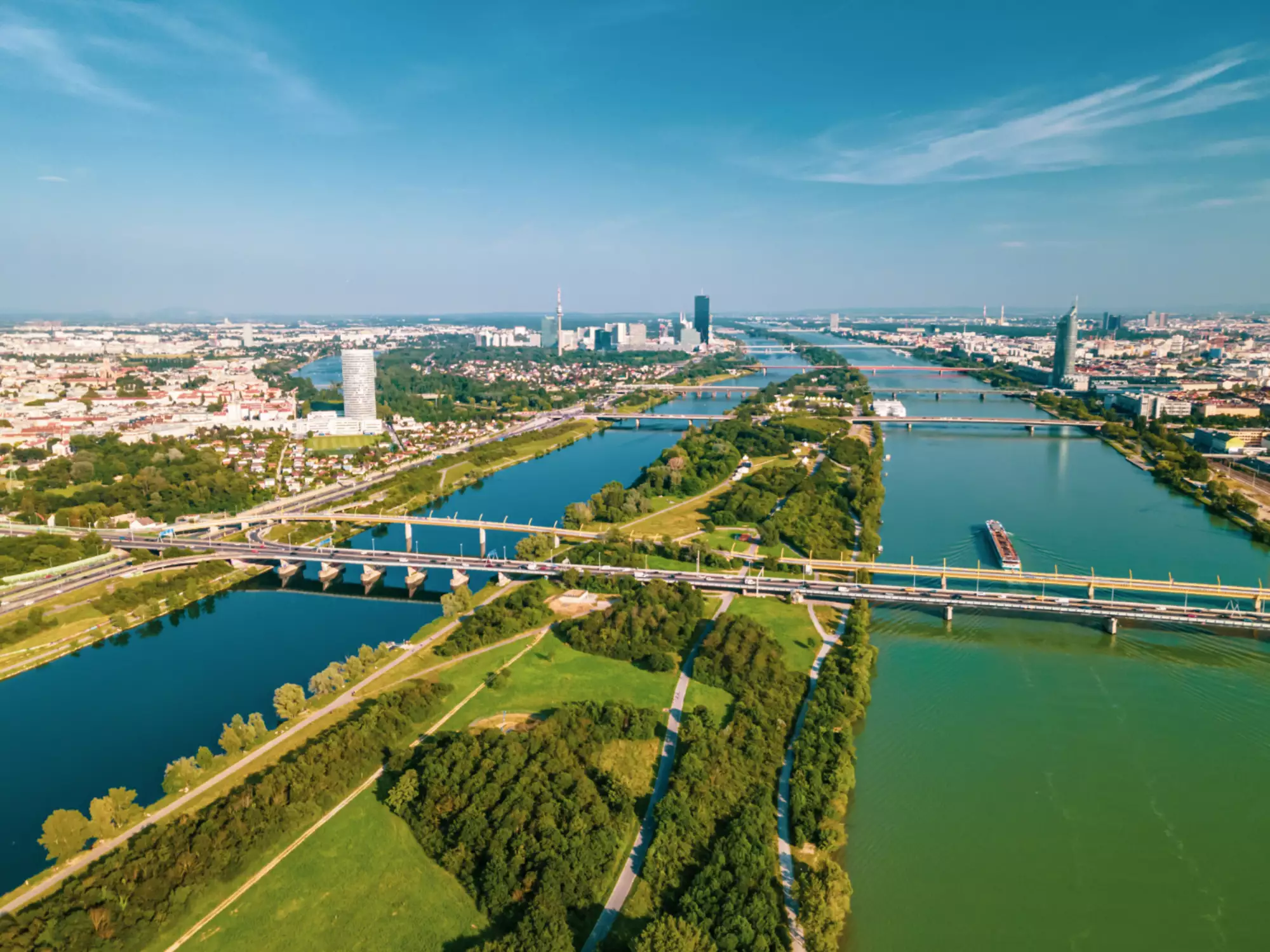 Luftaufnahme eines urbanen Gebiets mit Flüssen und Brücken. Grüne Parkflächen und Bäume säumen die Ufer. Im Hintergrund sind moderne Gebäude und eine Stadtlandschaft sichtbar, während der Himmel klar und blau ist.