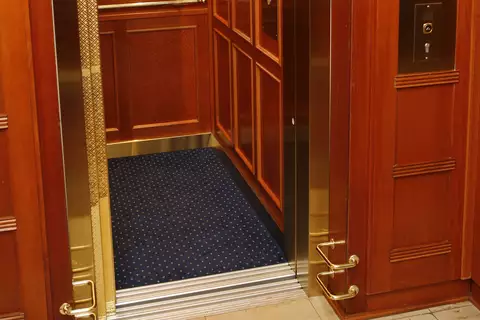 Ein eleganter Aufzug mit Holzwänden und goldenen Akzenten. Die Türen sind geöffnet, und der Boden ist mit einem blauen Teppich ausgelegt. An der Wand sind einige Knöpfe und Informationsblätter angebracht.