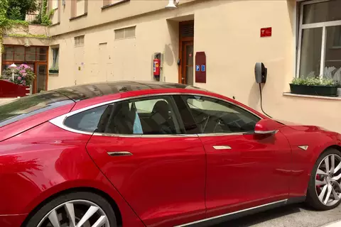 Ein rotes Elektroauto parkt neben der Wand eines Gebäudes mit einer kleinen Pflanze auf der Fensterbank. An der Wand sind zwei Ladestationen für Elektrofahrzeuge angebracht. Im Hintergrund ist ein Schild mit der Aufschrift "Hotel Wien" zu sehen.