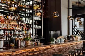 Eine stilvolle Bar mit einer modernen Theke aus Holz und vielen Sitzplätzen. Im Hintergrund steht ein Regal voller alkoholischer Getränke, beleuchtet von einigen hängenden Lampen. Die Atmosphäre wirkt einladend und elegant.