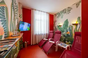 Der Raum zeigt eine entspannende Atmosphäre mit zwei roten Liegen und einer dekorativen Wand aus grünen Pflanzenmotiven. Ein Fernseher ist an der Wand befestigt, und ein Tisch mit Zeitschriften ist vorhanden. Der Raum hat große Fenster mit Vorhängen, die natürliches Licht hereinlassen.