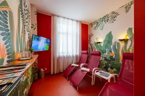 Der Raum zeigt eine entspannende Atmosphäre mit zwei roten Liegen und einer dekorativen Wand aus grünen Pflanzenmotiven. Ein Fernseher ist an der Wand befestigt, und ein Tisch mit Zeitschriften ist vorhanden. Der Raum hat große Fenster mit Vorhängen, die natürliches Licht hereinlassen.