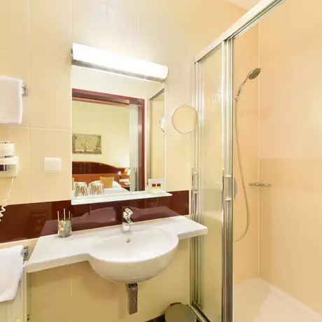 Das Bild zeigt ein modernes Badezimmer mit Dusche, Waschbecken und Spiegel. An der Wand hängen zwei Handtücher. An der Seite befindet sich ein Fön. Die Wände sind hell beige mit dunklen roten Akzenten.