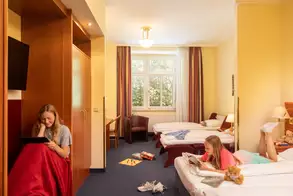 Zwei Mädchen in einem Hotelzimmer. Eines sitzt auf einem Stuhl mit einem Tablet, das andere liegt auf einem Bett und liest ein Buch. Der Raum hat zwei Betten und Fenster mit Tageslicht, der Boden ist unordentlich mit Spielzeug.