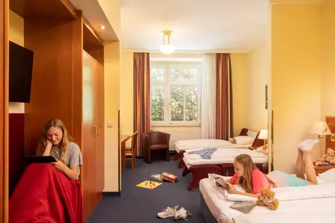 Zwei Mädchen in einem Hotelzimmer. Eines sitzt auf einem Stuhl mit einem Tablet, das andere liegt auf einem Bett und liest ein Buch. Der Raum hat zwei Betten und Fenster mit Tageslicht, der Boden ist unordentlich mit Spielzeug.