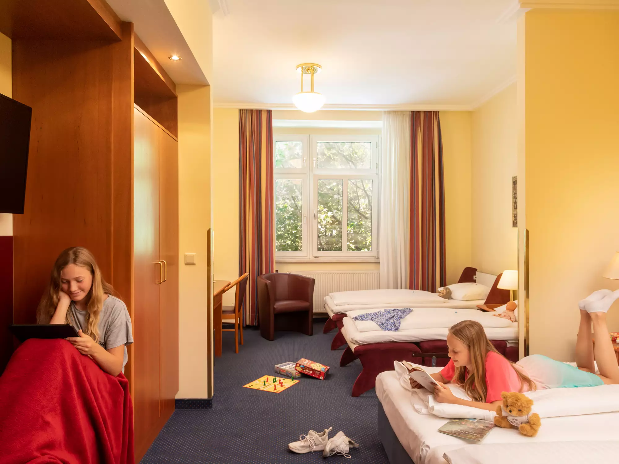 Zwei Mädchen in einem Hotelzimmer. Eines sitzt auf einem Stuhl mit einem Tablet, das andere liegt auf einem Bett und liest ein Buch. Der Raum hat zwei Betten und Fenster mit Tageslicht, der Boden ist unordentlich mit Spielzeug.