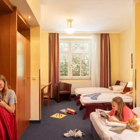 Zwei Mädchen in einem Hotelzimmer. Eines sitzt auf einem Stuhl mit einem Tablet, das andere liegt auf einem Bett und liest ein Buch. Der Raum hat zwei Betten und Fenster mit Tageslicht, der Boden ist unordentlich mit Spielzeug.