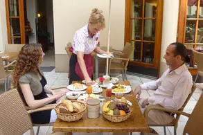 Eine Kellnerin serviert das Frühstück an einem Tisch im Freien. Zwei Gäste, eine Frau mit lockigem Haar und ein Mann in einem Hemd, sitzen auf Stühlen. Auf dem Tisch sind Teller mit Essen, wie Brot, Früchte und Getränke, sowie eine Obstschale zu sehen.