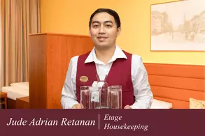 Ein Mitarbeiter im Housekeeping trägt zwei Wasserkaraffen und steht in einem Hotelzimmer. Im Hintergrund sind Möbelelemente sichtbar. Der Mitarbeiter ist freundlich und trägt eine Dienstkleidung.