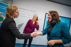 Drei Frauen stehen in einem Raum des Bundesministeriums. Zwei Frauen schütteln sich die Hände, während eine dritte im Hintergrund zuschaut. Alle tragen formelle Kleidung und wirken freundlich. An der Wand sind Informationen über Klimaschutz und Umwelt zu sehen.
