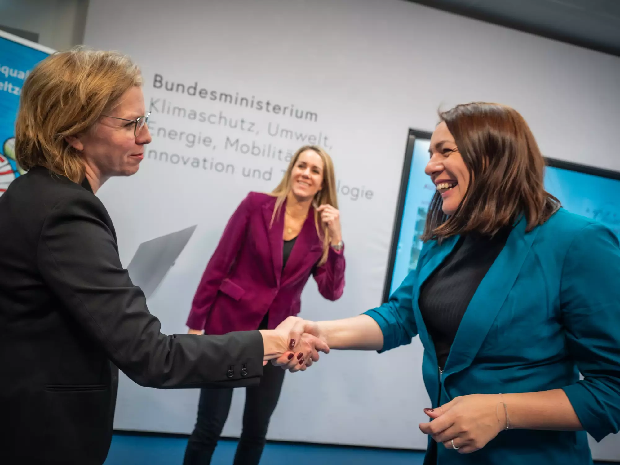 Drei Frauen stehen in einem Raum des Bundesministeriums. Zwei Frauen schütteln sich die Hände, während eine dritte im Hintergrund zuschaut. Alle tragen formelle Kleidung und wirken freundlich. An der Wand sind Informationen über Klimaschutz und Umwelt zu sehen.