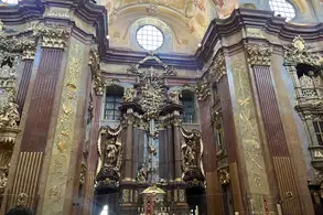 Der Innenraum einer kunstvoll dekorierten Kirche mit einer prunkvollen Altarwand, verziert mit Goldverzierungen und religiösen Figuren. Fenster und eine dekorierte Kuppel sind sichtbar, die ein feierliches Licht hereinlassen. Der Raum vermittelt eine Atmosphäre von Ehrfurcht und Pracht.
