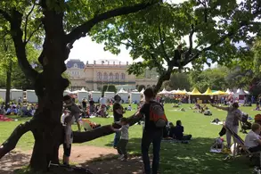 Ein belebter Park mit vielen Menschen, darunter Kinder, die auf einer Wiese spielen. Im Vordergrund steht ein großer Baum, an dem einige Kinder klettern. Im Hintergrund sind Festzelte und eine historische Architektur sichtbar. Es ist ein sonniger Tag.
