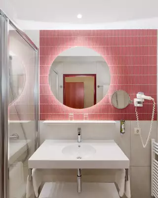 Ein modernes Badezimmer mit weißen Fliesen, einem Waschbecken auf einem schmalen Unterschrank, einem runden Spiegel und einer roten Wand mit einem kreisförmigen Muster. Es gibt eine Duschecke aus Glas, Handtuchhalter und einen Fön.