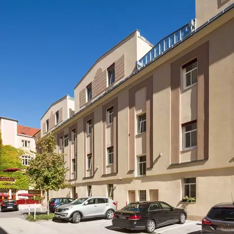 Ein Innenhof mit mehreren Gebäuden ist zu sehen. Die Wände sind hell gestrichen, und es sind verschiedene Autos geparkt. Im Hintergrund steht ein Gebäude mit grüner Fassade und dem Schriftzug "Hotel Wien". Der Himmel ist klar und blau.