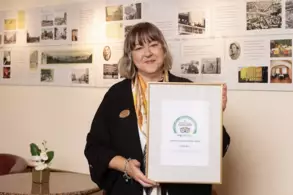 Eine Frau mit langer, lockiger Haarfrisur hält ein gerahmtes Zertifikat in der Hand, während sie vor einer Wand mit historischen Fotografien steht. Sie trägt eine schwarze Jacke und ein buntes Tuch um den Hals.