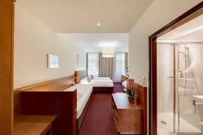 Ein Blick auf ein Hotelzimmer mit zwei Einzelbetten. Die Wände sind hell, und große Fenster lassen Tageslicht herein. Ein Holzschrank und ein Fernseher sind sichtbar. Rechts ist ein Badezimmer mit Dusche zu sehen. Ein paar Pflanzen dekorieren den Raum.