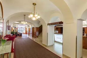 Ein hell erleuchteter Empfangsbereich eines Hotels mit Bögen und einer roten Teppichfläche. Links stehen Pflanzen und Möbel, während sich rechts eine Empfangstheke mit der Beschriftung "RECEPTION" befindet. Eine Treppe führt zu einem oberen Bereich. Große Fenster bieten Sicht nach außen.