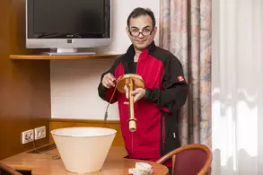 Ein Mann in einem schwarzen und roten Outfit steht an einem Tisch in einem Hotelzimmer. Er hält ein Lampenteilen in der Hand und schaut zur Kamera. Im Hintergrund sind eine Fernseher und Vorhänge zu sehen. Der Tisch hat eine große, weiße Schüssel und einige Materialien.