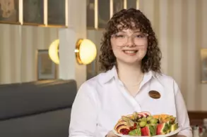 Eine lächelnde Person trägt einen Teller mit einer bunten Auswahl an frischem Obst. Im Hintergrund sind schemenhaft dekorative Wände zu sehen. Die Person trägt eine weiße Bluse und eine Namensplakette.