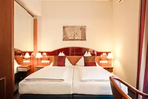 Ein gemütliches Hotelzimmer mit zwei Einzelbetten, die mit weißen Bettwäsche und roten Kissen dekoriert sind. An den Wänden sind Holzverkleidungen und ein Spiegel. Auf den Nachttischen stehen kleine Lampen. Ein Kunstwerk hängt über den Betten.