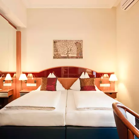 Ein gemütliches Hotelzimmer mit zwei Einzelbetten, die mit weißen Bettwäsche und roten Kissen dekoriert sind. An den Wänden sind Holzverkleidungen und ein Spiegel. Auf den Nachttischen stehen kleine Lampen. Ein Kunstwerk hängt über den Betten.