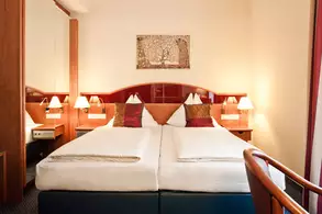 Ein gemütliches Hotelzimmer mit zwei Einzelbetten, die mit weißen Bettwäsche und roten Kissen dekoriert sind. An den Wänden sind Holzverkleidungen und ein Spiegel. Auf den Nachttischen stehen kleine Lampen. Ein Kunstwerk hängt über den Betten.