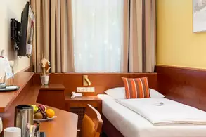 Das Bild zeigt ein gemütliches Hotelzimmer mit einem Einzelbett, einem Tisch und einem Stuhl. An der Wand hängt ein Fernseher, und auf dem Tisch stehen ein Wasserkocher sowie frisches Obst. Vor einem Fenster sind Vorhänge sichtbar.