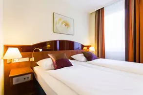 Das Bild zeigt ein gemütliches Hotelzimmer mit zwei Einzelbetten, die mit weißer Bettwäsche und dekorativen Kissen in Lila ausgestattet sind. An den Wänden hängen Vorhänge in warmen Farbtönen, und die Beleuchtung kommt von zwei Nachttischlampen. Ein Bild mit einer Blume ist an der Wand angebracht.