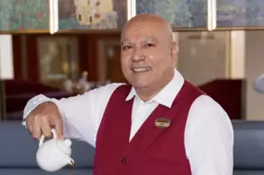 Ein Herr in einer roten Weste lächelt, während er aus einer Teekanne in eine Teetasse gießt. Im Hintergrund sind stilvolle Restaurantmöbel und dekorative Kunstwerke zu sehen.