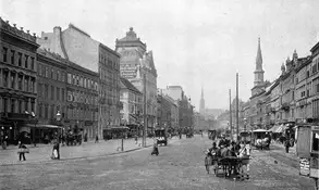 Eine historische Stadtansicht mit einer breiten Straßenmitte, umgeben von Gebäuden im architektonischen Stil des späten 19. Jahrhunderts. In der Ferne sind Kirchtürme sichtbar, während Menschen und Fuhrwerke die Straße beleben. Die Szene wirkt nostalgisch und vermittelt den Charme einer vergangenen Epoche.