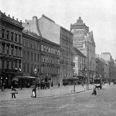 Eine historische Stadtansicht mit einer breiten Straßenmitte, umgeben von Gebäuden im architektonischen Stil des späten 19. Jahrhunderts. In der Ferne sind Kirchtürme sichtbar, während Menschen und Fuhrwerke die Straße beleben. Die Szene wirkt nostalgisch und vermittelt den Charme einer vergangenen Epoche.