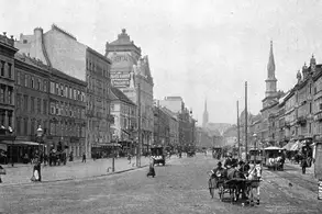 Eine historische Stadtansicht mit einer breiten Straßenmitte, umgeben von Gebäuden im architektonischen Stil des späten 19. Jahrhunderts. In der Ferne sind Kirchtürme sichtbar, während Menschen und Fuhrwerke die Straße beleben. Die Szene wirkt nostalgisch und vermittelt den Charme einer vergangenen Epoche.