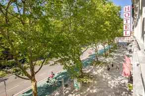 Ein Straßenszenario mit einem großen Hotelzeichen „HOTEL WIEN“ und einer lebhaften Umgebung. Zwei Radfahrer fahren auf einem Radweg, während Fußgänger auf dem Gehweg unterwegs sind. Umgeben von grünen Bäumen und sonnigem Wetter.