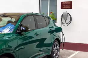 A green electric vehicle is parked near a wall with an electric charging station and a plaque above it. The car is plugged in, indicating it is charging, with a clear view of the vehicle's sleek design and shiny surface.