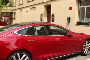 Ein rotes Elektroauto parkt neben der Wand eines Gebäudes mit einer kleinen Pflanze auf der Fensterbank. An der Wand sind zwei Ladestationen für Elektrofahrzeuge angebracht. Im Hintergrund ist ein Schild mit der Aufschrift "Hotel Wien" zu sehen.