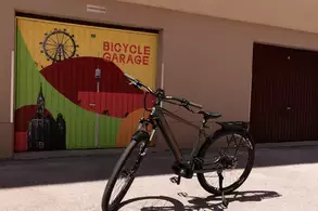Ein Fahrrad steht auf einem gepflasterten Boden vor einer Garage mit buntem Garagentor, das die Aufschrift "BICYCLE GARAGE" trägt. Im Hintergrund sind stilisierte Stadtansichten und ein Riesenrad zu sehen.