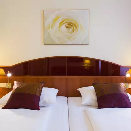 Das Bild zeigt ein stilvoll eingerichtetes Hotelzimmer mit zwei Einzelbetten, die mit weißen Bettwäsche und roten Kissen ausgestattet sind. An der Wand hängt ein Bild einer weißen Rose. Es gibt zwei Nachttische mit Lampen und einem Telefon.
