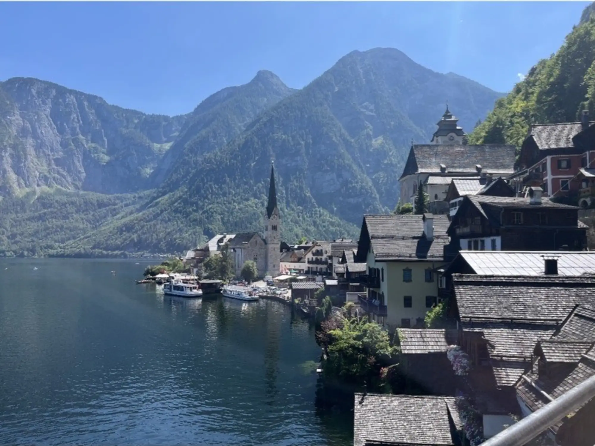 Blick auf das malerische Dorf Hallstatt, umgeben von hohen Bergen und einem klaren See. Die charmanten Holzhäuser liegen am Ufer, und der Kirchturm ragt in den Himmel. Klarer blauer Himmel und grünes Umland tragen zur wunderschönen Landschaft bei.