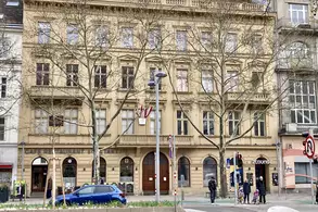 Eine historische, dreistöckige Gebäudeansicht mit großen Fenstern. Vor dem Gebäude stehen mehrere Bäume und einige Fußgänger. Ein blauer PKW parkt am Straßenrand. Im Vordergrund sind Blumen und Straßenelemente zu sehen.