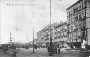 Eine historische Schwarz-Weiß-Aufnahme der Praterstraße in Wien, mit Gebäuden im Hintergrund, Straßenbahnen und Fußgängern. Ein hoher Laternenmast und eine Straßenlaterne sind sichtbar. Die Szene vermittelt einen Eindruck des städtischen Lebens zu Beginn des 20. Jahrhunderts.