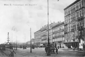 Eine historische Schwarz-Weiß-Aufnahme der Praterstraße in Wien, mit Gebäuden im Hintergrund, Straßenbahnen und Fußgängern. Ein hoher Laternenmast und eine Straßenlaterne sind sichtbar. Die Szene vermittelt einen Eindruck des städtischen Lebens zu Beginn des 20. Jahrhunderts.