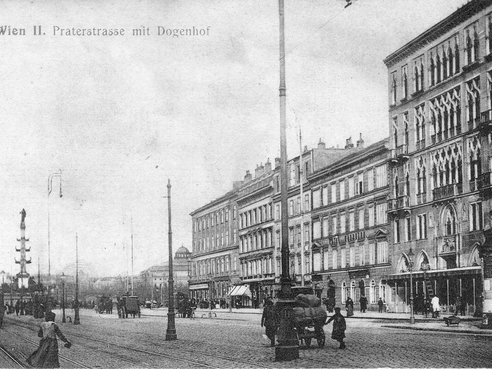 Eine historische Schwarz-Weiß-Aufnahme der Praterstraße in Wien, mit Gebäuden im Hintergrund, Straßenbahnen und Fußgängern. Ein hoher Laternenmast und eine Straßenlaterne sind sichtbar. Die Szene vermittelt einen Eindruck des städtischen Lebens zu Beginn des 20. Jahrhunderts.