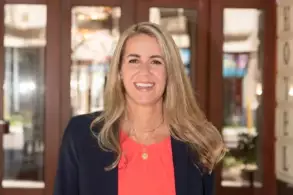 Eine Frau mit langen, blonden Haaren steht vor einer Eingangstür eines Hotels. Sie trägt ein rotes Oberteil und einen dunklen Blazer. Die Frau lächelt freundlich in die Kamera. Im Hintergrund sind Pflanzen und die Konturen des Hotelgebäudes sichtbar.