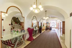 Der Empfangsbereich eines Hotels mit einer eleganten Einrichtung. Links ist eine Glasvitrine mit Blumen und rechts eine rote Tischdecke. Über dem Boden hängen runde Lichter, und der Boden ist mit einem roten Teppich ausgelegt. Im Hintergrund sind große Fenster zu sehen.