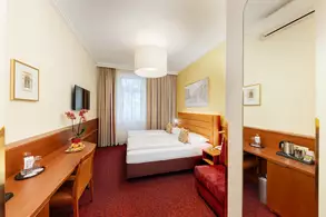 Ein gemütliches Hotelzimmer mit zwei Einzelbetten, einem großen Fenster mit Vorhängen und einem Kunstwerk an der Wand. Ein Schreibtisch mit Stuhl, Obstschale und einem Wasserkocher sind ebenfalls vorhanden. Der Boden ist mit rotem Teppich ausgelegt und die Wände sind in sanften Farben gehalten.